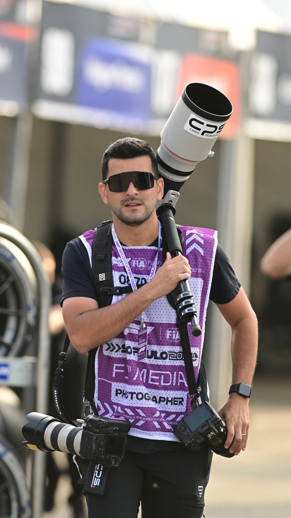 Lucas Miranda caminhando no paddock com teleobjetiva no ombro e colete de fotógrafo da F1